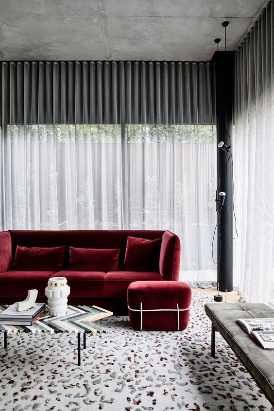 a refined living room with concrete walls, sheer curtains, a deep red modern sofa and a footrest, a chevron coffee table and a grey upholstered bench