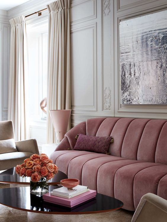 a refined living room with molding, a dusty pink sofa, a duo of black coffee tables, a creamy chair, some blooms and some books