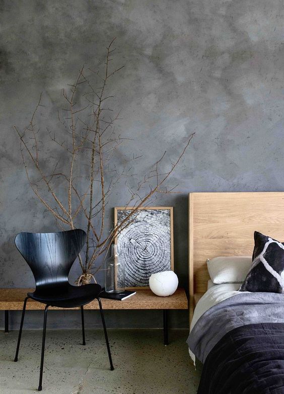 a refined modern bedroom with grey limewashed walls, a light-stained bed with dark bedding, a cork bench and a black plywood chair, some branches
