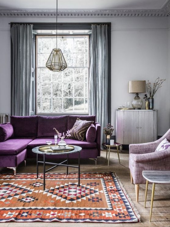 a serene living room with light grey walls, grey curtains, a purple sectional, a lilac chair and a printed rug
