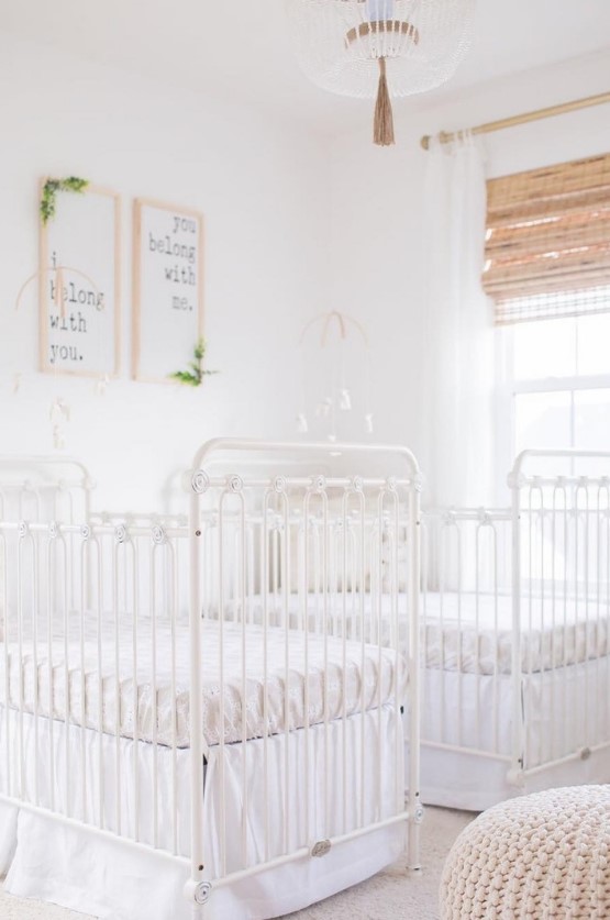 a serene white nursery with vintage white cribs, a knit ottoman, shades, artworks and a pretty chandelier plus matching mobiles
