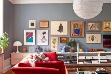 a slate blue living room with a catchy geometric red sofa, a glass table on casters, a large colorful gallery wall and an open storage unit with books