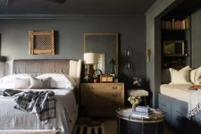 a slate grey bedroom with a cozy reading nook with built-in shelves, a creamy bed with neutral bedding, stained nightstands, layered rugs