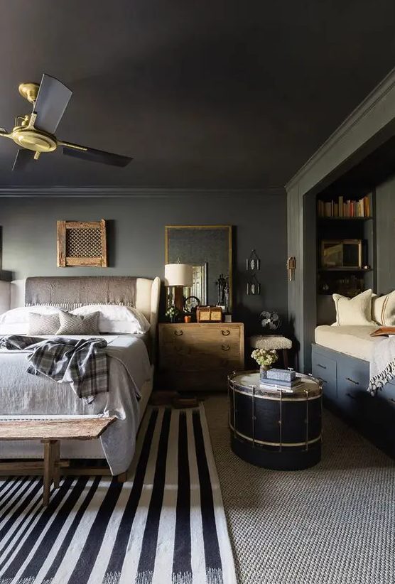 a slate grey bedroom with a cozy reading nook with built-in shelves, a creamy bed with neutral bedding, stained nightstands, layered rugs