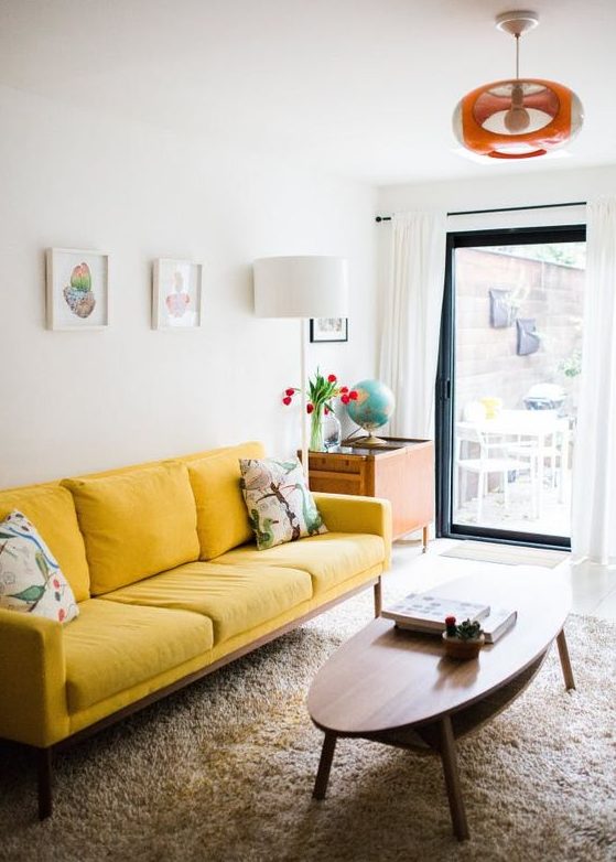 a small and cheerful living room with a yellow sofa, a low oval table, a small gallery wall and a bright lamp