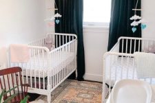 a small and cute twin nursery with white cribs, navy black-out curtains, a printed rug, potted plants and a lovely retro lamp