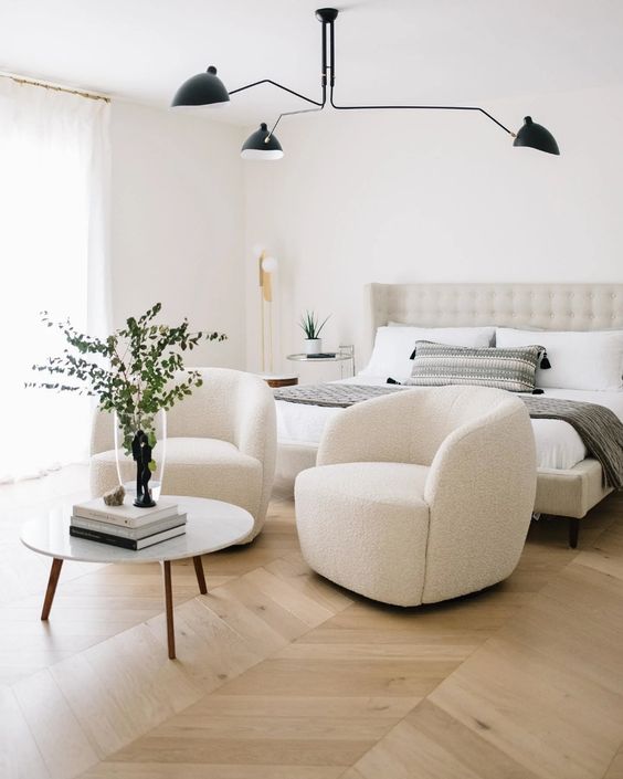 a sophisticated neutral bedroom with an upholstered bed, matching creamy chairs and a round table, a black chandelier and some lamps