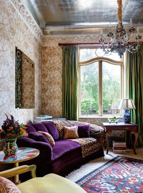 a sophisticated vintage living room with printed wallpaper, a purple sofa, a lovely chandelier, green curtais and a printed rug