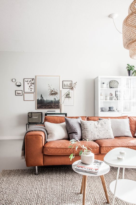 a stylish Scandinavian living room with an orange leather sofa and neutral pillows, a glass storage cabinet, a gallery wall and side tables