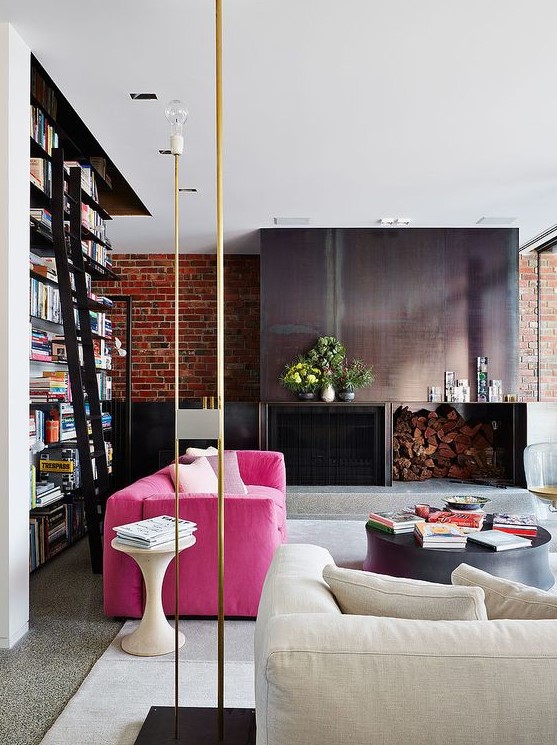 a stylish contemporary living room with a metal fireplace and firewood storage, a hot pink sofa, a neutral one, a large bookcase and gold touches