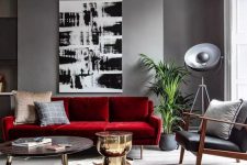 a stylish grey living room with a deep red sofa, a round table and a gold side table, a black leather chaur, catchy decor and potted plants