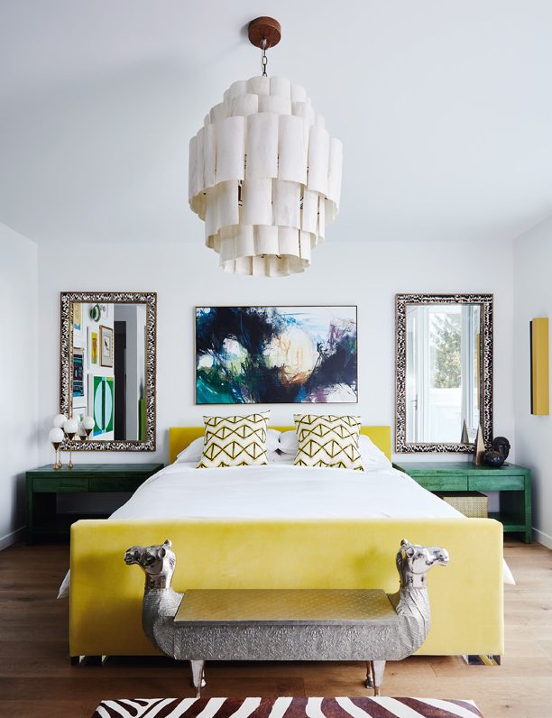 a stylish maximalist bedroom done in white, with a yellow upholstered bed, green nightstands, mirrors, a statement artwork and a catchy chandelier