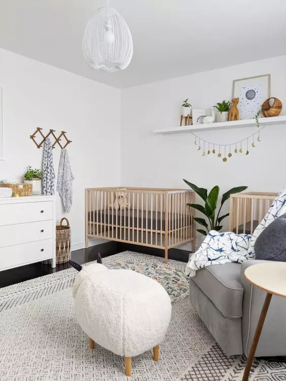 a stylish neutral twin nursery with stained cribs, a white dresser as a changing table, an open shelf with decor, a grey chair, an animal pouf and layered rugs