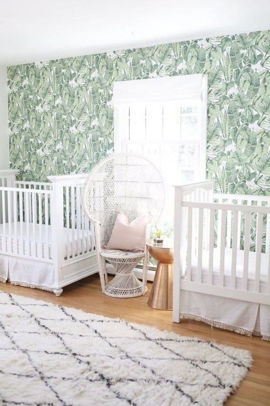 a tropical twin nursery with a banana leaf wall, white cribs and a peacock chair, a printed rug and a metallic side table