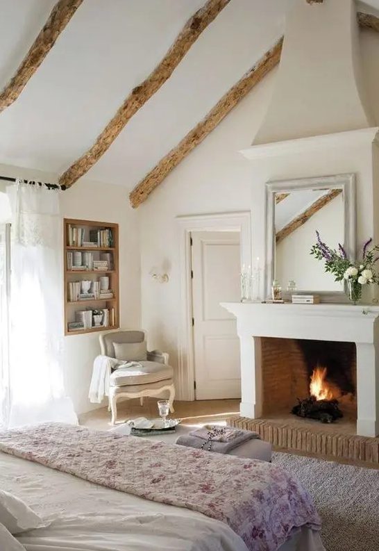 a vintage farmhouse bedroom in white and with wooden beams, with a fireplace, printed textiles