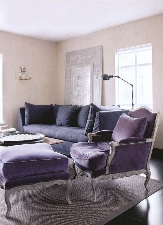 a vintage-inspired living room with tan walls, a deep purple sofa, a purple vintage chair and a footrest, a coffee table and a black floor lamp