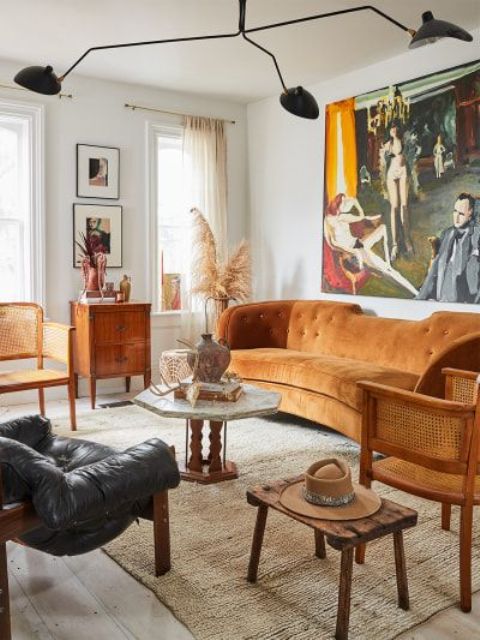 a vintage living room with an oversized artwork, a rust colored curved sofa, a black leather chair, cane chairs and a black chandelier