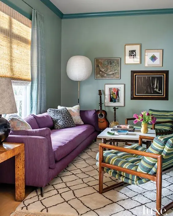 a welcoming living room with aqua walls, a purple sofa with printed pillows, a coffee table and stiped chairs, a gallery wall