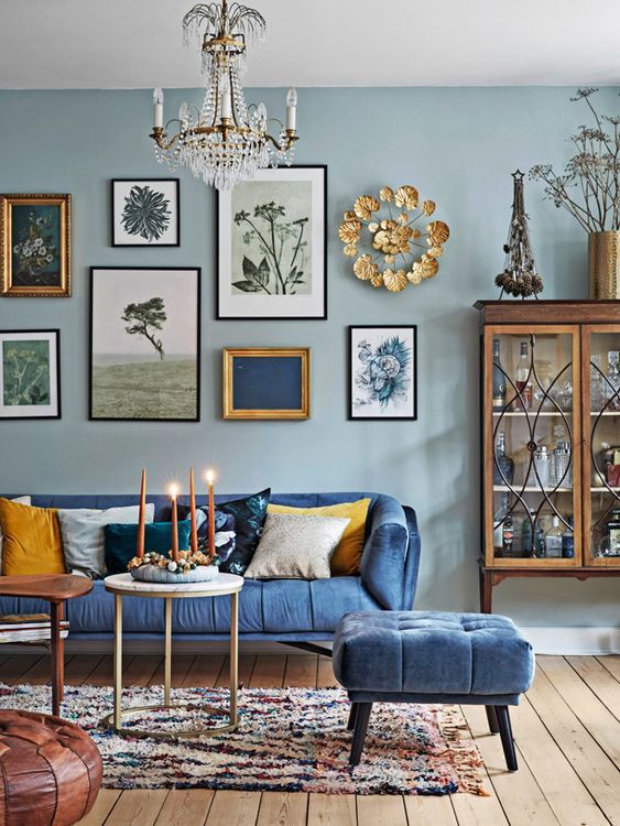 a welcoming living room with light blue walls, a blue sofa and a pouf, mismatching coffee tables, a gallery wall and a vintage glass cabinet
