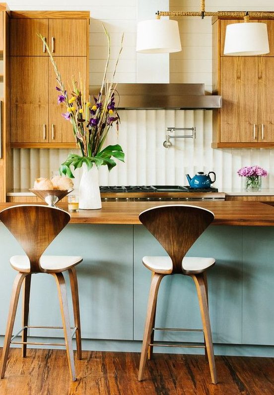 a welcoming mid century modern kitchen with light colored cabinets with metallic handles, plywood stools, a blue kitchen island and a wooden countertop