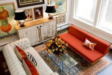 a whimsical living room with a bold gallery wall, a white tufted sofa, an orange sofa, a glass table and a shabby chic cabinet