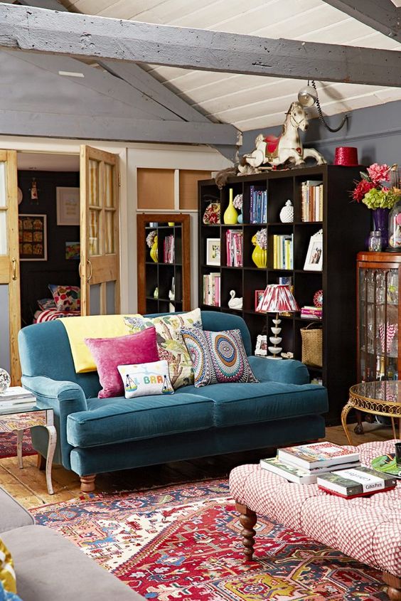 an eclectic living room with a blue sofa, a printed rug, colorful pillows, an ottoman with books, a dark-stained bookcase and bright decor