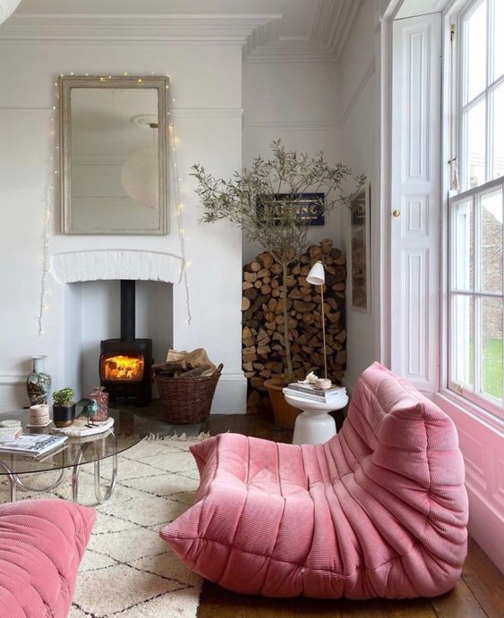 an eclectic living room with a hearth, pink loveseat, a glass table, firewood and side tables, potted trees and plants