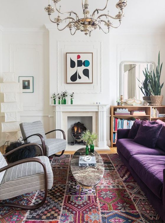 an eclectic living room with a non-working fireplace, a depe purple sofa, printed chairs, a refined chandelier and a bold printed rug