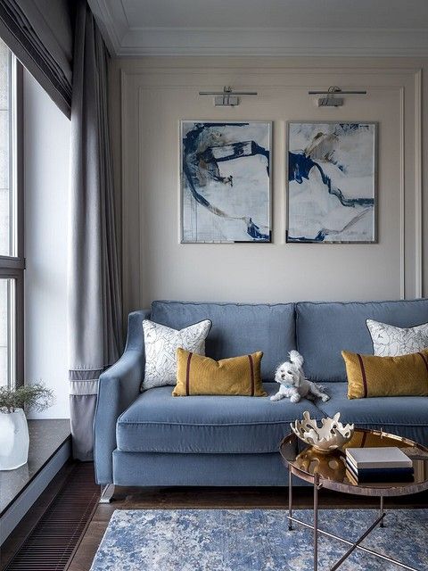 an elegant and classic living room with a light blue sofa, a shiny coffee table, watercolor artworks, grey curtains, a blue rug and cool pillows