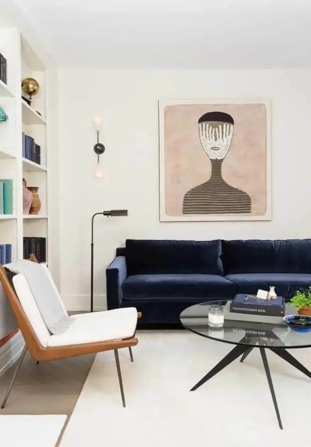 an elegant creamy living room with open storage units, a midnight blue velvet sofa, a chic chair, a bold artwork, a round coffee table and some greenery