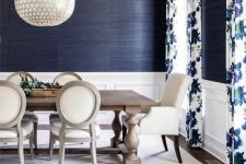 an elegant dining room with navy grasscloth wallpaper, white paneling, a stained dining table and neutral chairs, a pendant lamp