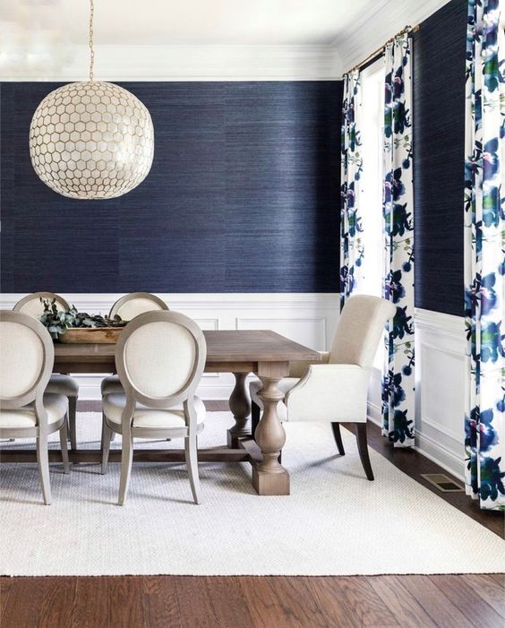 an elegant dining room with navy grasscloth wallpaper, white paneling, a stained dining table and neutral chairs, a pendant lamp
