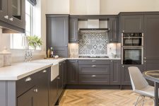 an elegant kitchen with slate grey shaker style cabinets and white stone countertops, a printed tile backspalsh and grey and white furniture