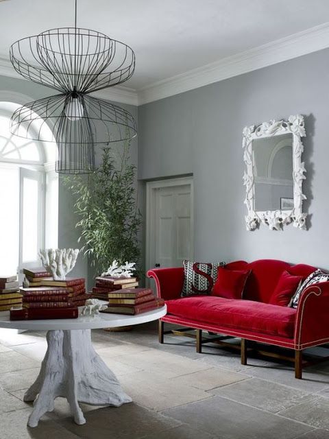 an elegant living room with light grey walls, a deep red velvet sofa, a large round table, a wire chandelier and a mirror in an ornated frame