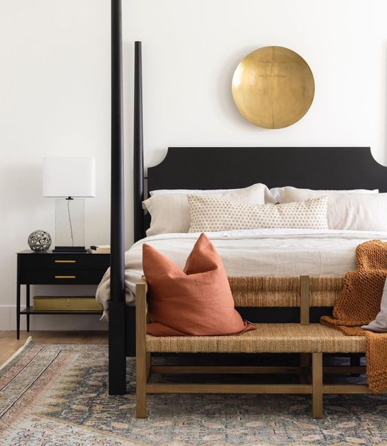 an elegant modern bedroom with a black bed with pillars, black nightstands, a woven bench with coral pillows and a rust blanket