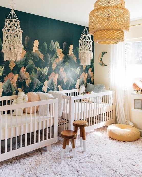 an eye-catchy boho twin nursery with a tropical leaf accent wall, white cribs, wooden stools, macrame and a pendant lamp is wow