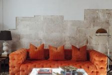 an industrial living room with rough walls, a refined orange tufted sofa, a low glass table and floor lamps