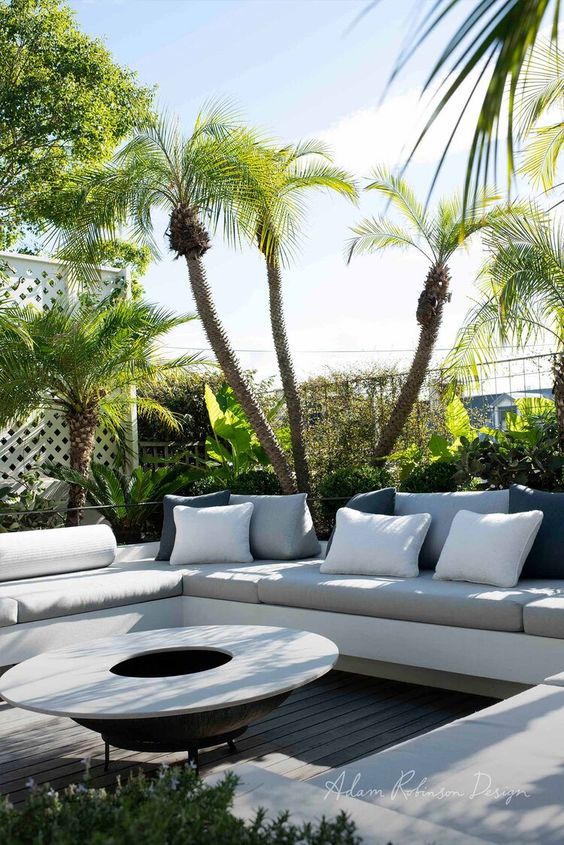 a contemporary terrace with a built-in sofa with neutral upholstery, a portable fire pit and lots of tropical plants around