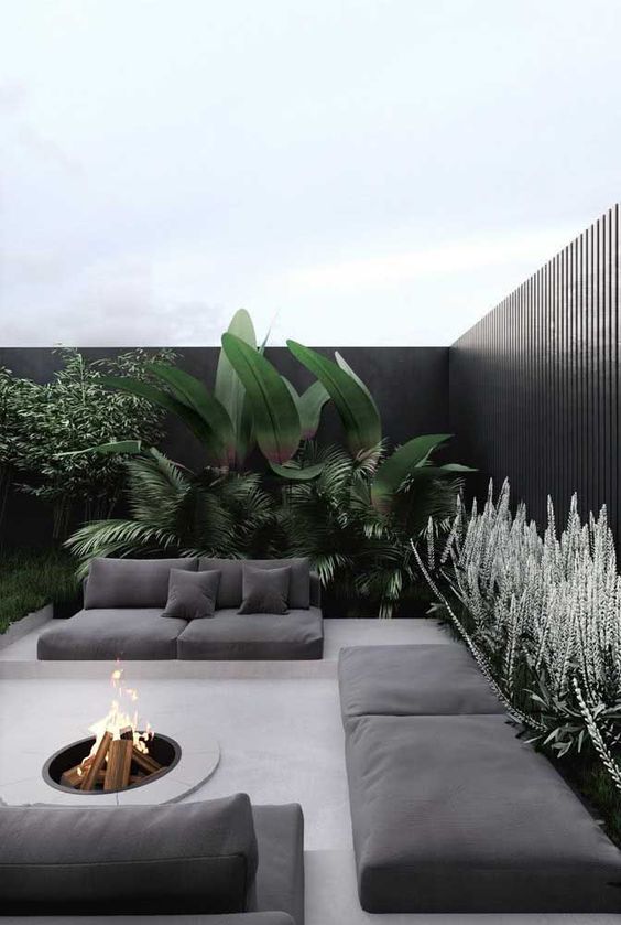 a minimalist tropical space with lots of plants, a tall black fence, built-in concrete sofas and a daybed with graphite grey cushions and a fire pit