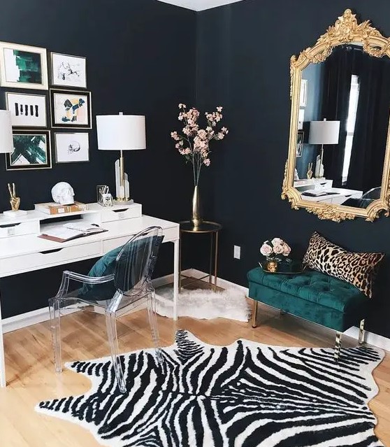 a refined working space with black walls, a white desk, an acrylic chairs, a dark green upholstered ottoman, a mirror in a gilded frame, a zebra print rug and a leopard print pillow