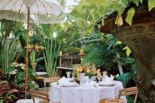 09 a small tropical terrace with a dining space, with folding chairs and a round table, a carved storage unit and candles, an umbrella and a tropical garden around