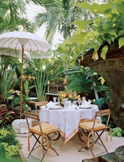 a small tropical terrace with a dining space, with folding chairs and a round table, a carved storage unit and candles, an umbrella and a tropical garden around