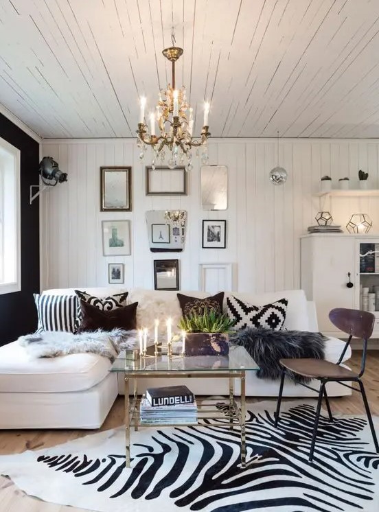 a Scandinavian living room with a gallery wall, a crystal chandelier, a white corner sofa and printed pillows, a zebra print rug and a glass coffee table
