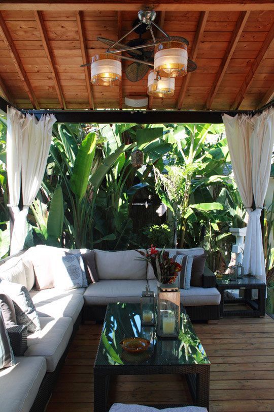 a tropical cabana surrounded with lush tropical plants, with white curtains, a neutral sectional and a black coffee table plus candle lanterns