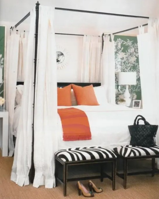 a stylish bedroom with green botanical print walls, a canopy bed with white curtains, orange and white pillows and zebra print stools at the foot of the bed