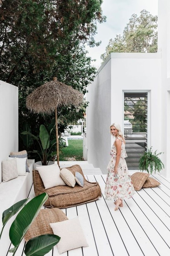a tropical porch with a whitewashed wooden floor, burlap furniture, an umbrella, potted greenery and pillows