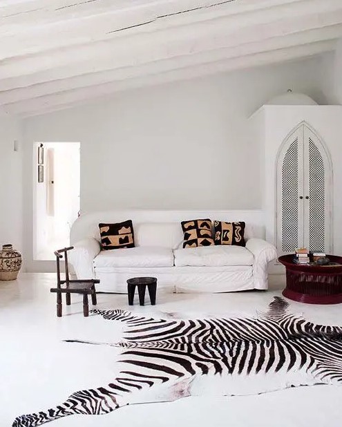 an eclectic white living room with a wardrobe, a white sofa, black stools, a mahogany coffee table and a zebra print rug plus printed pillows