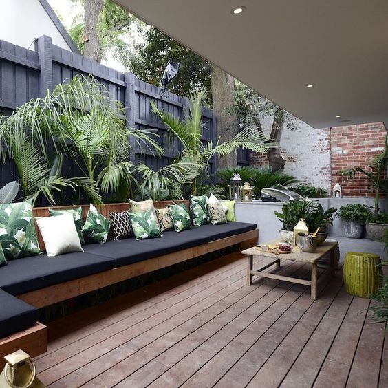 a contemporary outdoor space with a built-in wooden sofa, a wooden coffee table, leaf print pillows and lots of tropical plants around