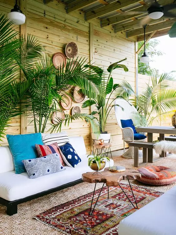 a boho tropical space with a wooden wall, a white sofa with colorful pillows, a living edge table and printed rugs