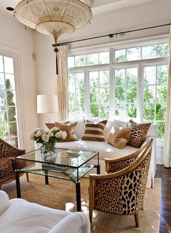 a refined living room with a white sofa, a glass tiered coffee table, cheetah printed chairs and a pillow, a jute rug and a beautiful pendant lamp